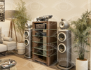 A high-end wooden audio equipment cabinet with glass shelves, holding a turntable and receiver, flanked by two matching floorstanding speakers in a styled living room setting.